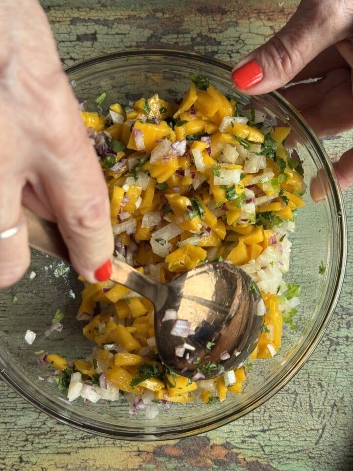 a hand stirring mango salsa in a glass bowl