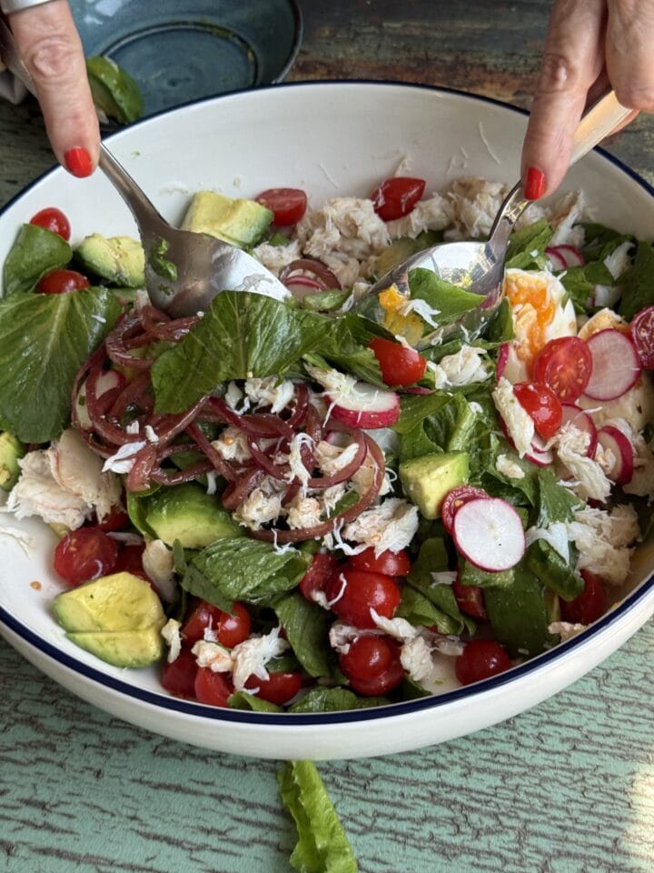 two hands with red nail polish tossing a salad in a big serving bowl