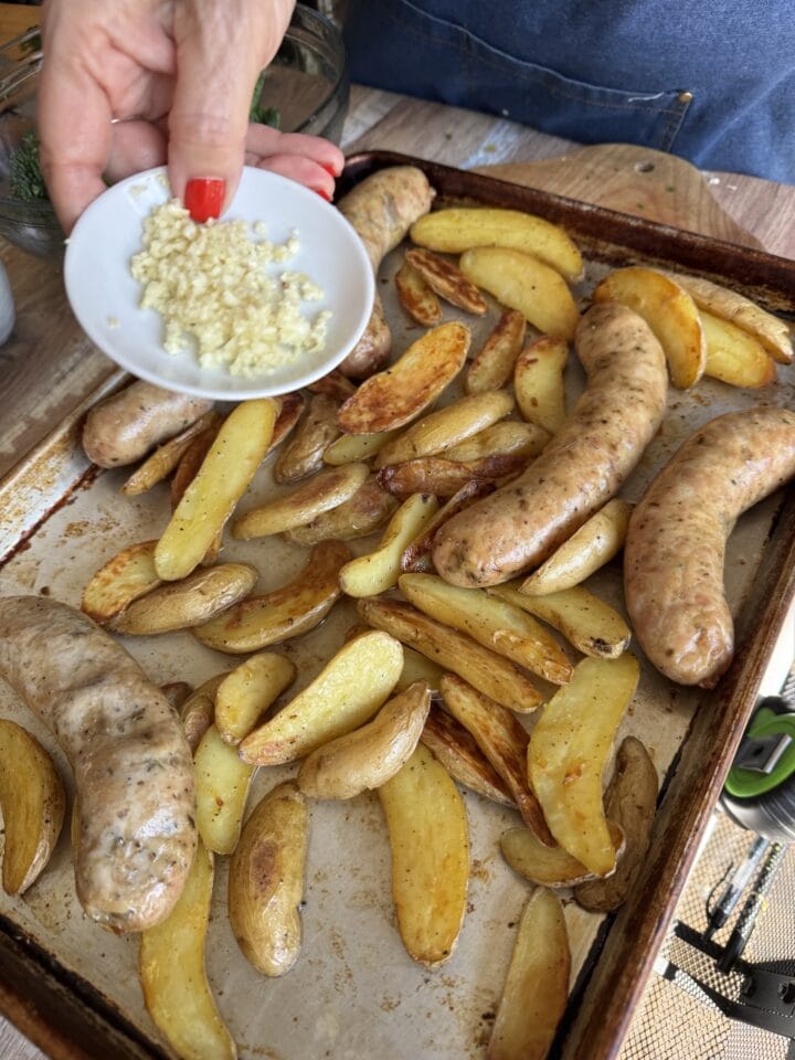 a small plate of chopped garlic above a sheet pan with sausage and potatoes