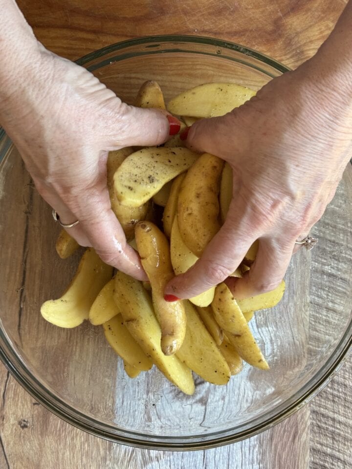 hands tossing fingerling potatoes in olive oil in a glass bowl