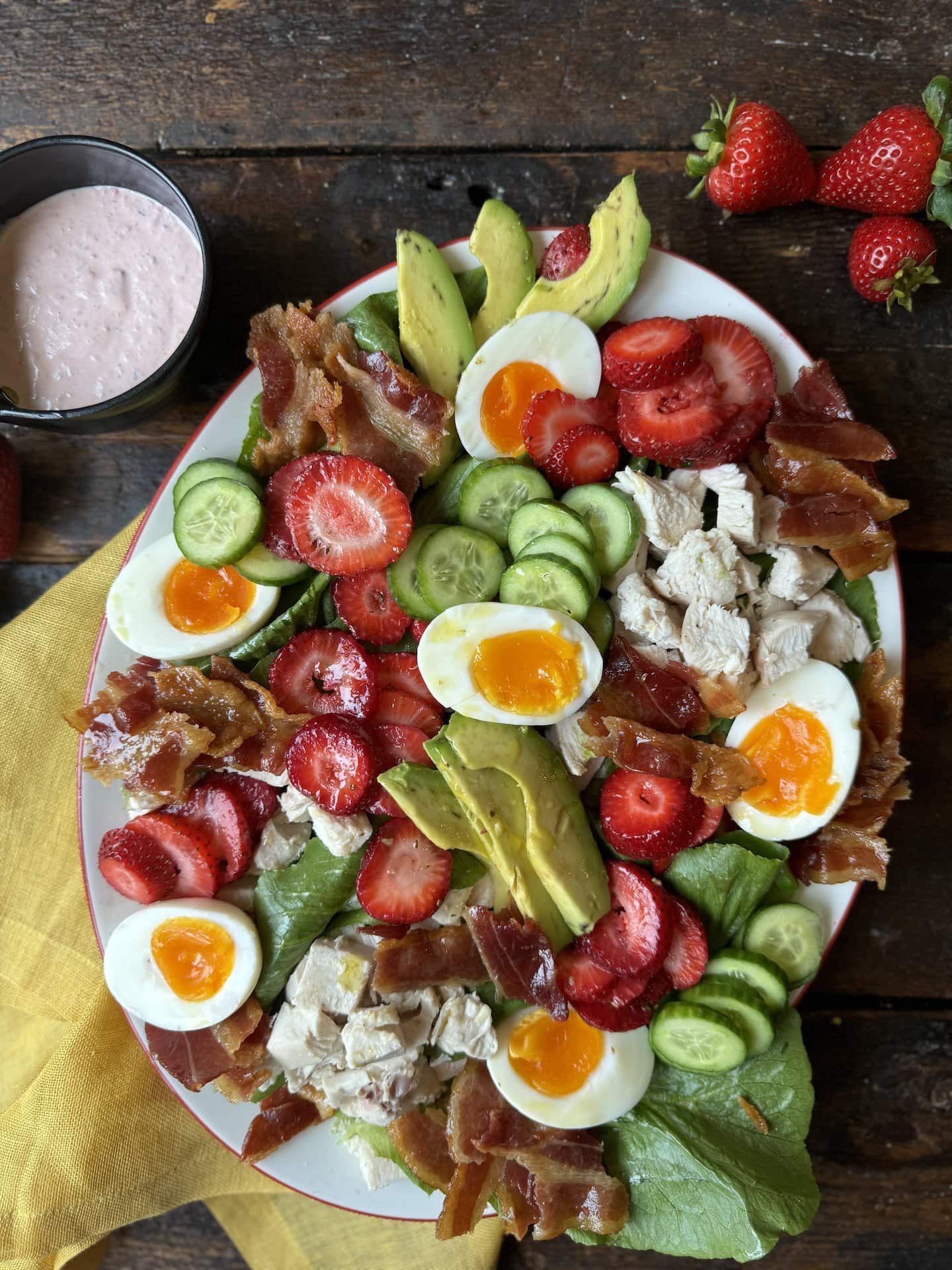 Strawberry Cobb Salad on platter next to strawberries and bowl with strawberry chive dressing on a yellow napkin