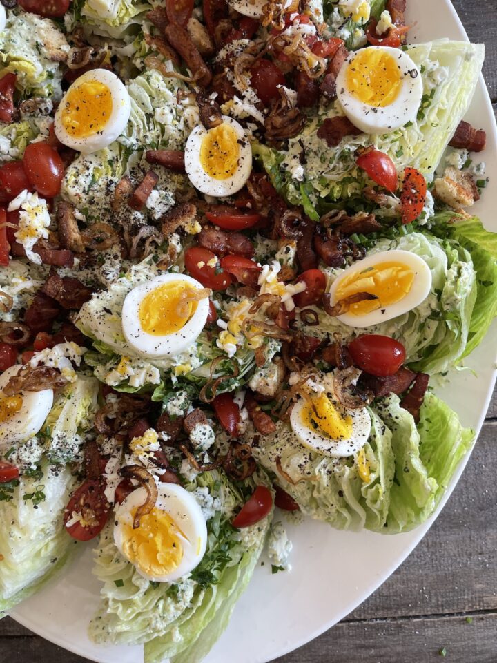 A classic wedge salad with wedges of iceberg lettuce, hard boiled eggs, bacon, cherry tomato, blue cheese, and chives.