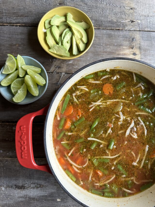 A dutch oven full of beef& noodle soup next to a bowl of limes and avocado slices.