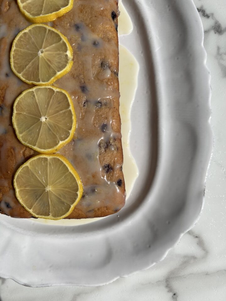 whole lemon blueberry cake topped with icing & lemon slices on on white platter 