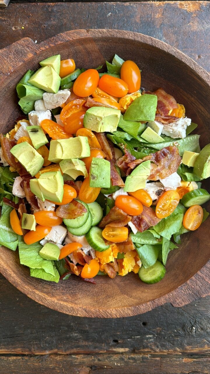 Fresh avocado and cherry tomato salad with bacon, chicken, and cucumbers in a wooden bowl.