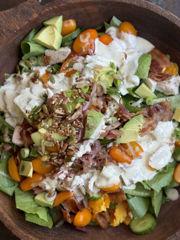 Fresh loaded salad with avocados, cherry tomatoes, bacon, boiled eggs, and creamy dressing in a wooden bowl.