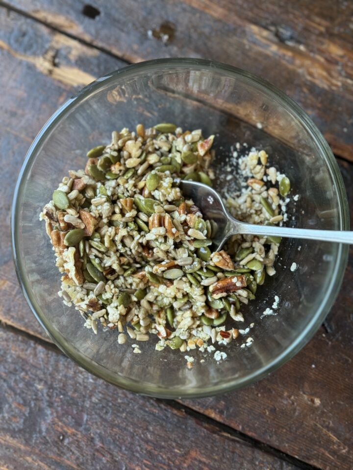 nuts and seeds in a glass bowl with a metal spoon on a wood counter