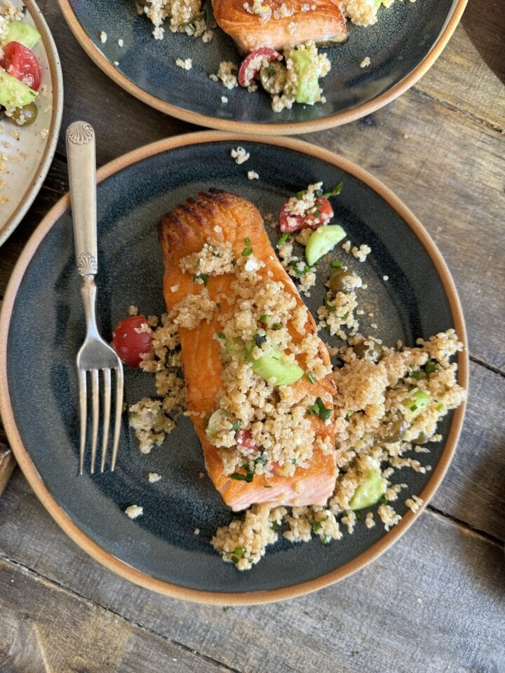 a photo of salmon on a blue plate with greek quinoa salad on top and a fork next to it