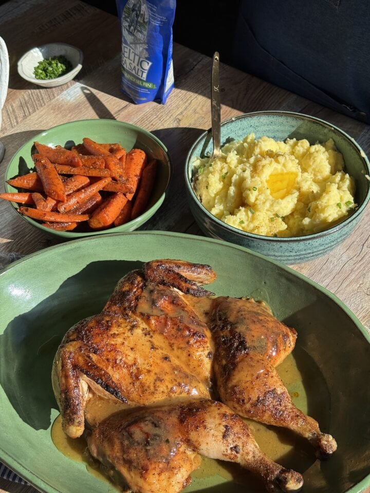 roasted spatchcock chicken on green platter next to two bowls with spiced carrots and buttery mashed potatoes and a small blue bag of salt in the background 