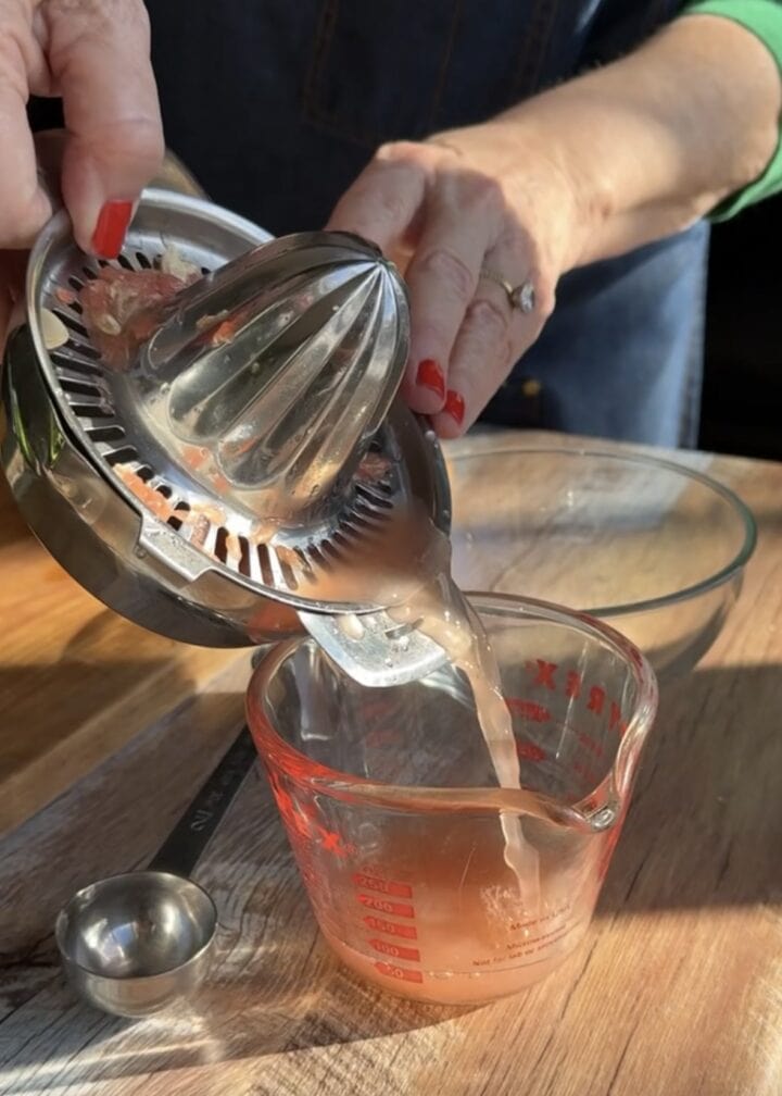 two hands with red nail polish pouring grapefruit juice from a metal juicer into a glass measuring cup 
