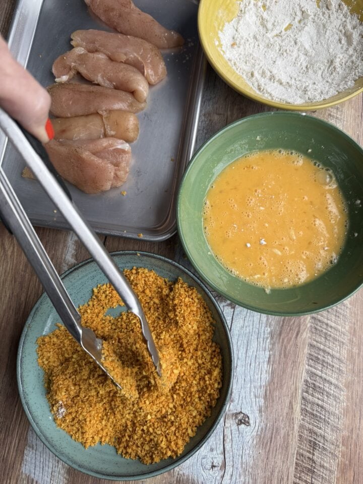 chicken tenderloins next to three bowls filled with flour, eggs, and breading