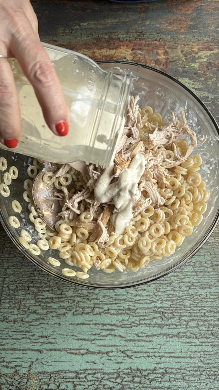 caesar dressing pouring into a glass bowl with pasta and chicken in it