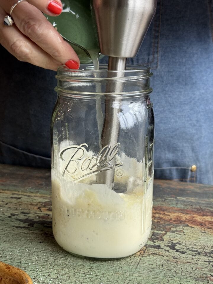 caesar dressing in a mason jar with an immersion blender in it and lemon juice pouring in