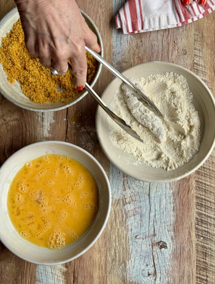 flour, breading, and eggs in small bowls with a chicken tenderloin in the flour