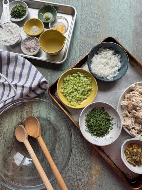 all ingredients for high protein chicken salad in small bowls next to a glass bowl with wooden spoons in it