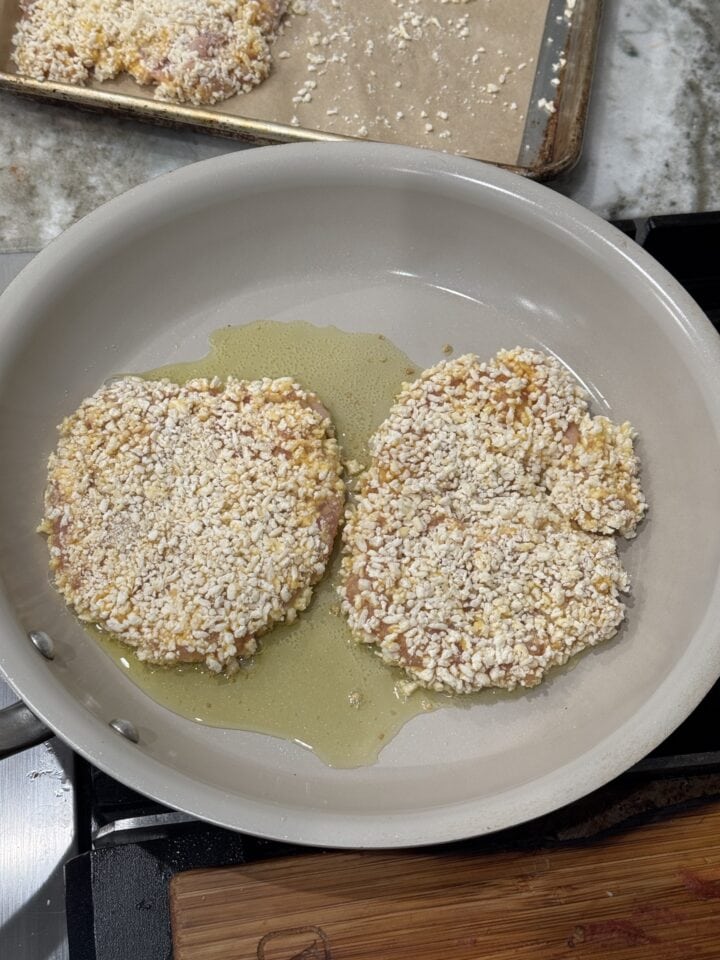 two breaded chicken cutlets cooking in oil in a pan
