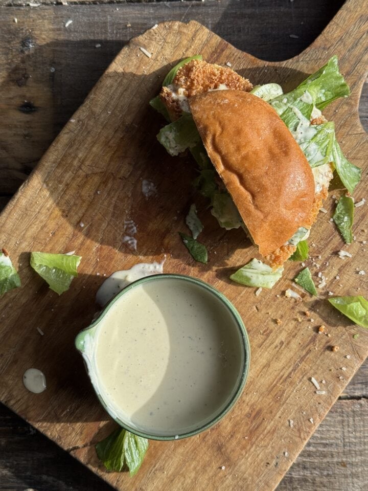 half of a sandwich on a wooden cutting board with a small bowl of dressing next to it