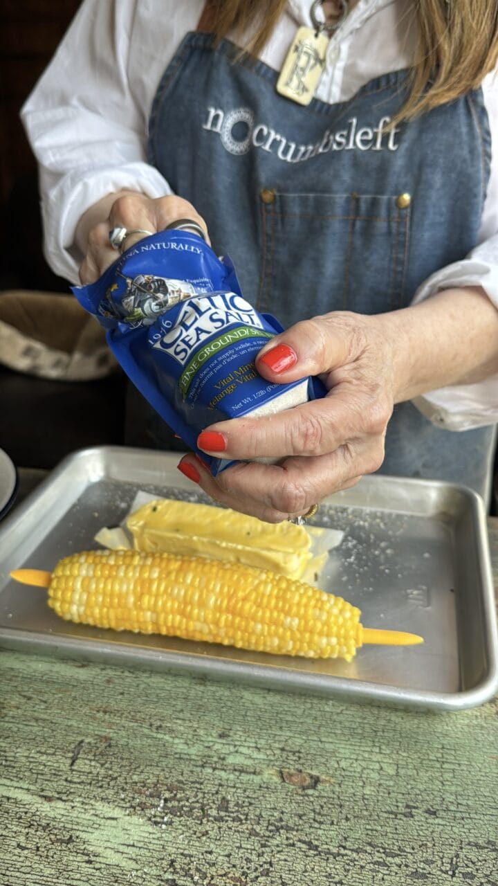 teri reaching in a bag of celtic sea salt above a butter corn cob