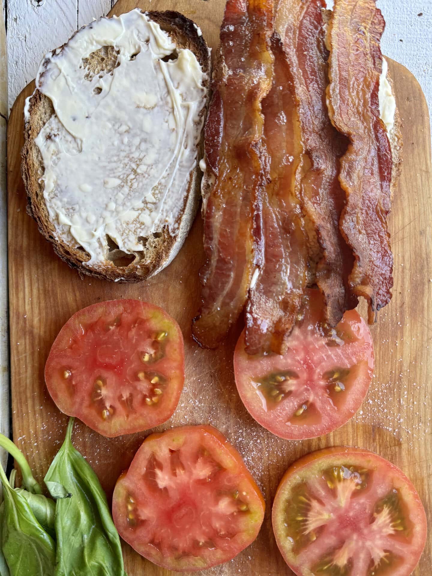 bread with mayo on one side and oven bacon on top of the other, with tomato slices next to it