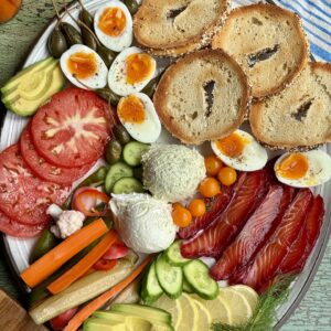 a platter with salt cured salmon, bagels, jammy eggs, and vegetables on it