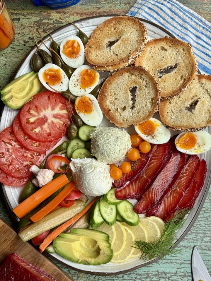 a platter with salt cured salmon, bagels, jammy eggs, and vegetables on it