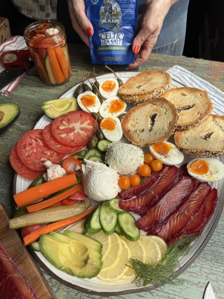 a platter with salt cured salmon, bagels, jammy eggs, and vegetables on it and pickles in the background